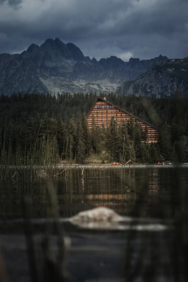 Skalnaté štíty pod těžkými mraky, pod nimi jehličnatý les, hotel s trojúhelníkovou střechou a tmavá hladina s rákosím.
