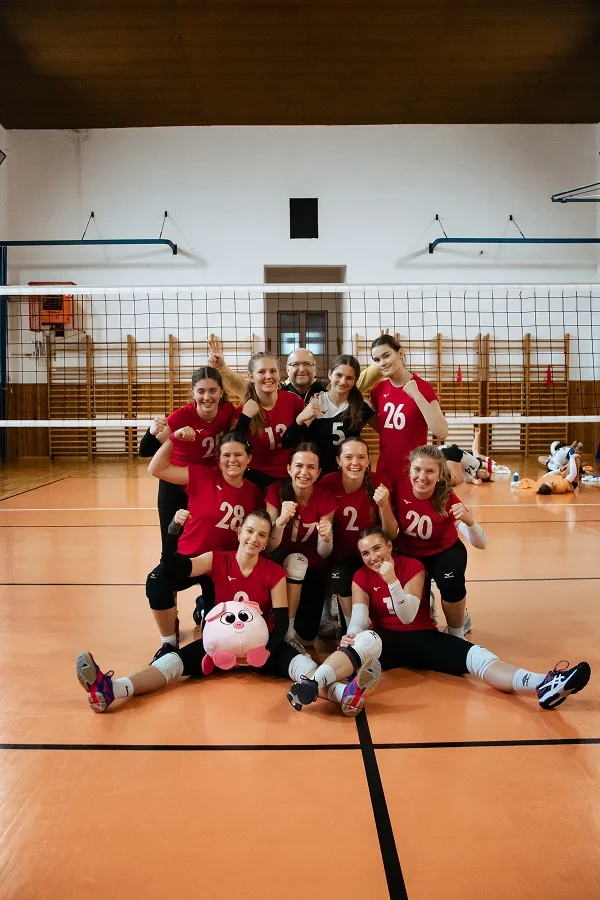 Usměvavé volejbalistky v červených dresech ukazují svaly v jasně osvětlené tělocvičně, vzadu muž v brýlích, vpředu plyšové prasátko.