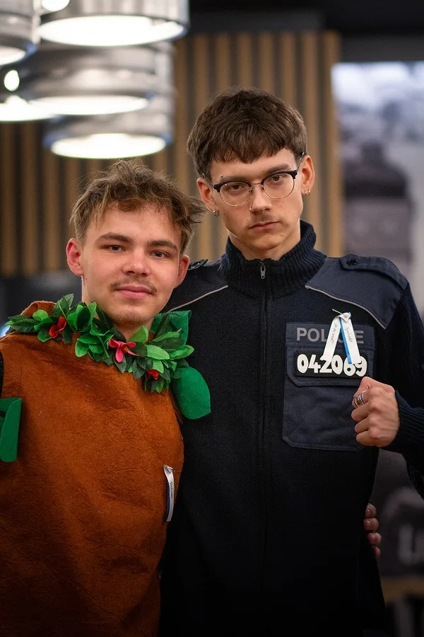 Dva mladíci v kostýmech pózují pod lampami: jeden s věncem z listí, druhý v policejní uniformě se stužkou.