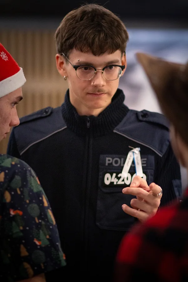 Mladík v brýlích a policejním kostýmu drží stužku připnutou na hrudi, v měkkém světle mezi rozostřenými spolužáky.